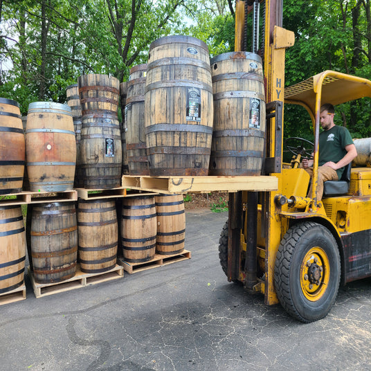 A man operates a forklift, lifting wooden barrels onto a stack on a paved surface in an outdoor industrial area.