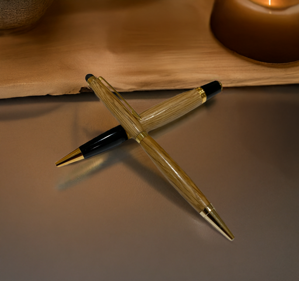 Two Barrel Pens, handcrafted from oak whiskey barrels and finished with gold accents, are crossed on a smooth surface. A candle and wooden tray sit in the background.