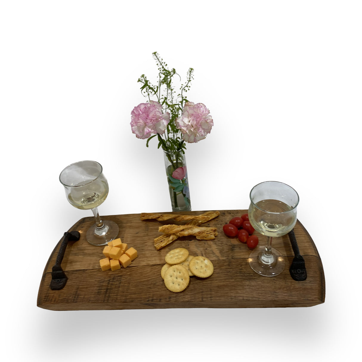 The Charcuterie and Cheese Rectangle Wooden Serving Board, full size and hand made from solid wood, holds wine, cheese, crackers, breadsticks, and cherry tomatoes next to a pink floral vase for rustic charm.