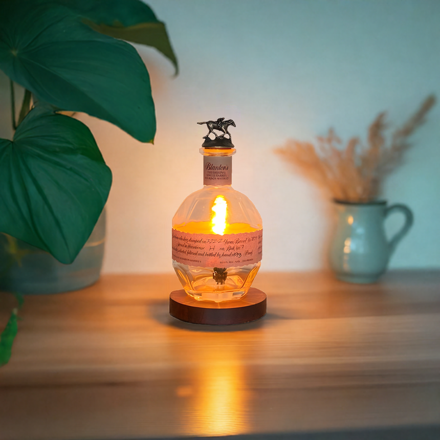 The Blanton's Lamp, with its warm glow, sits on a wooden surface near a plant and ceramic jug, bringing an elegant touch to the decor.