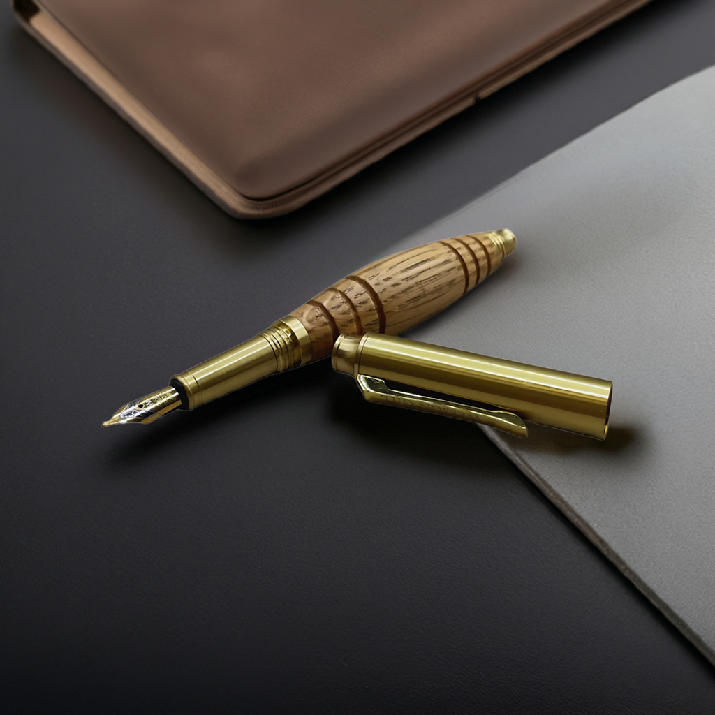 The Bourbon Barrel Fountain Pen, handcrafted from reclaimed Kentucky bourbon barrels with a gold and wood body, rests uncapped on a dark desk beside a notebook.