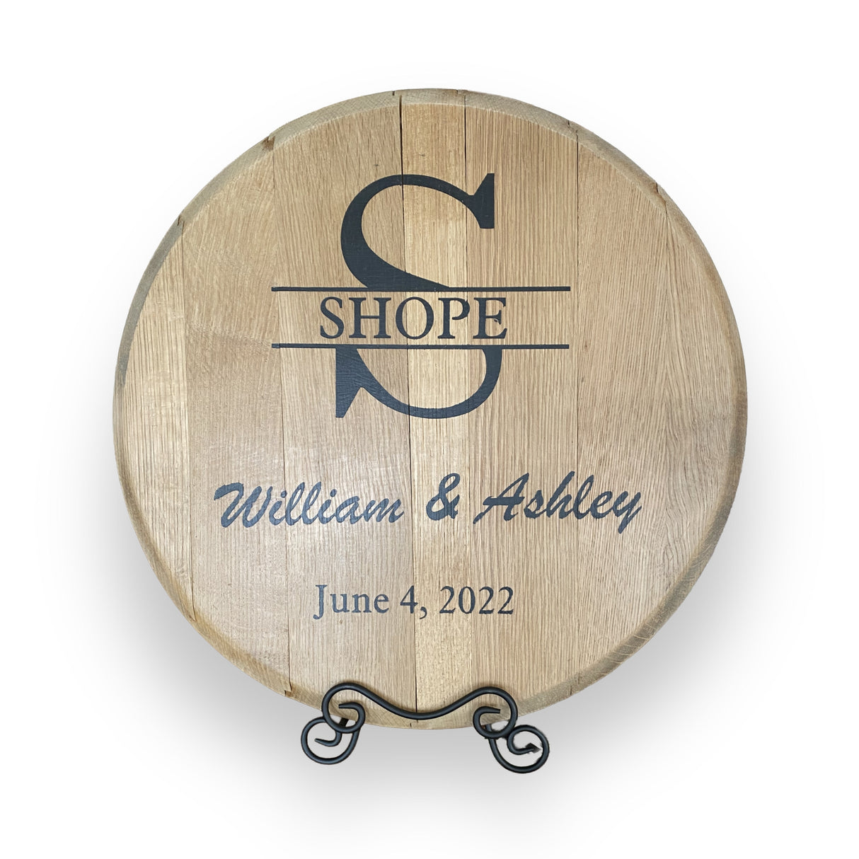 The Whiskey Barrel Head Wedding Special is a round wooden sign made from authentic barrel heads, engraved with "SHOPE," "William & Ashley," and a wedding date, displayed on a metal stand—an unforgettable personalized keepsake.