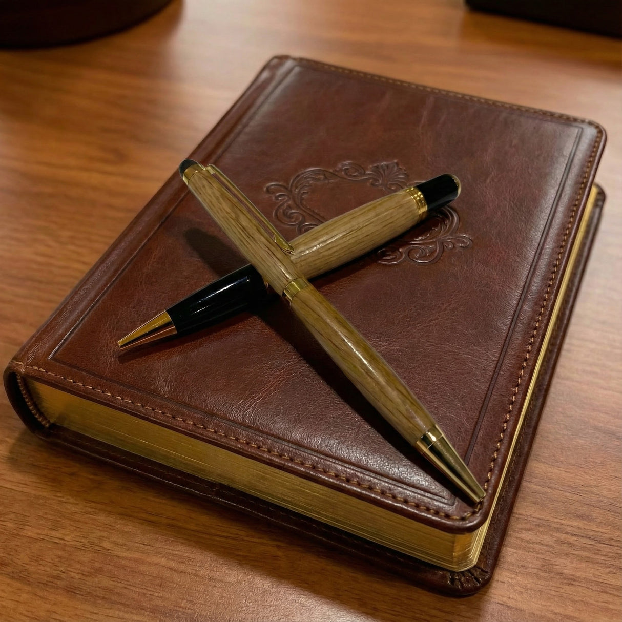 A Barrel Pen and another pen arranged in an X shape sit atop a brown leather-bound book with gold-edged pages.