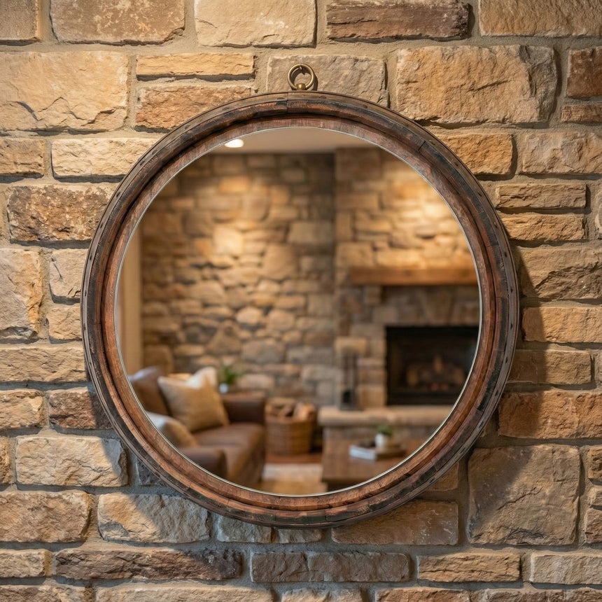 The Round Mirror, made from reclaimed whiskey barrels, hangs on a stone wall and reflects a cozy living room with a couch, fireplace, and soft lighting.