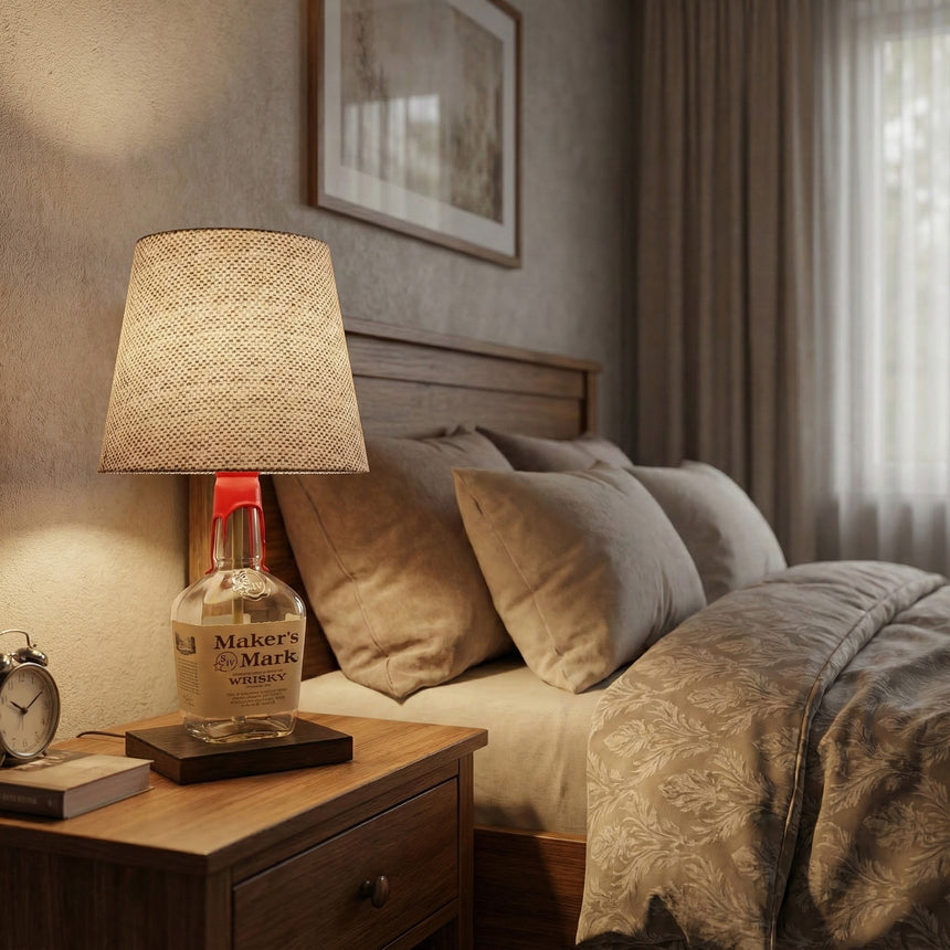 The Maker's Mark Lamp, crafted from a bourbon bottle, sits on a wooden nightstand beside a neatly made bed, adding distinctive charm to your home decor.