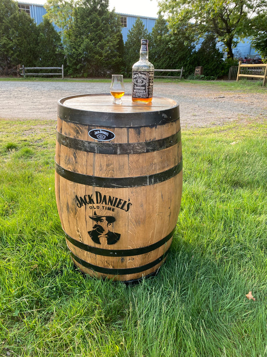 A Jack Daniel’s barrel with a whiskey bottle and glass on top, placed on grassy ground outside.