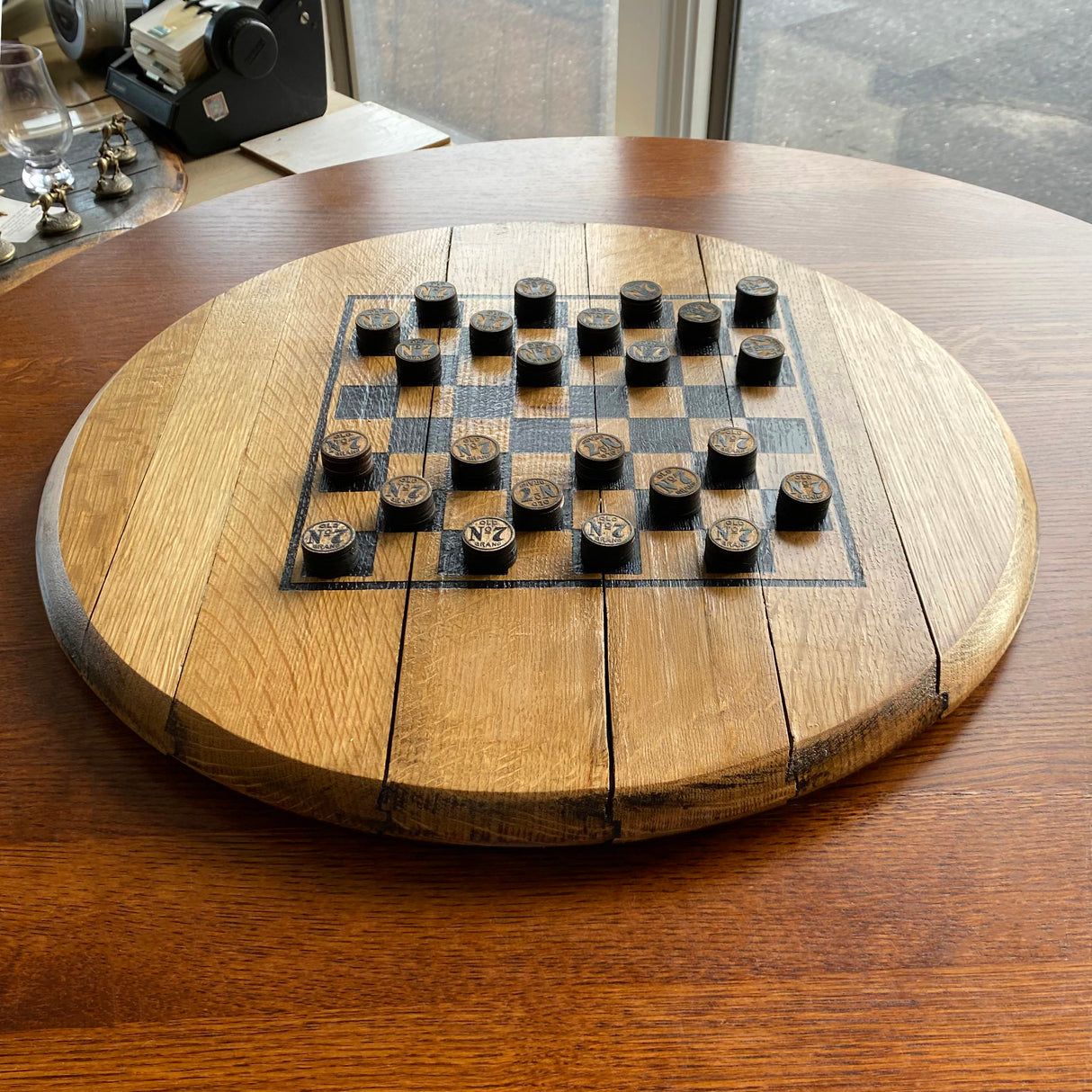 A Chess and Checkers Board With Solid Wood Barrel Head Base, featuring hand made playing pieces, rests on a table by the window with black and tan checkers set up for versatile gameplay.