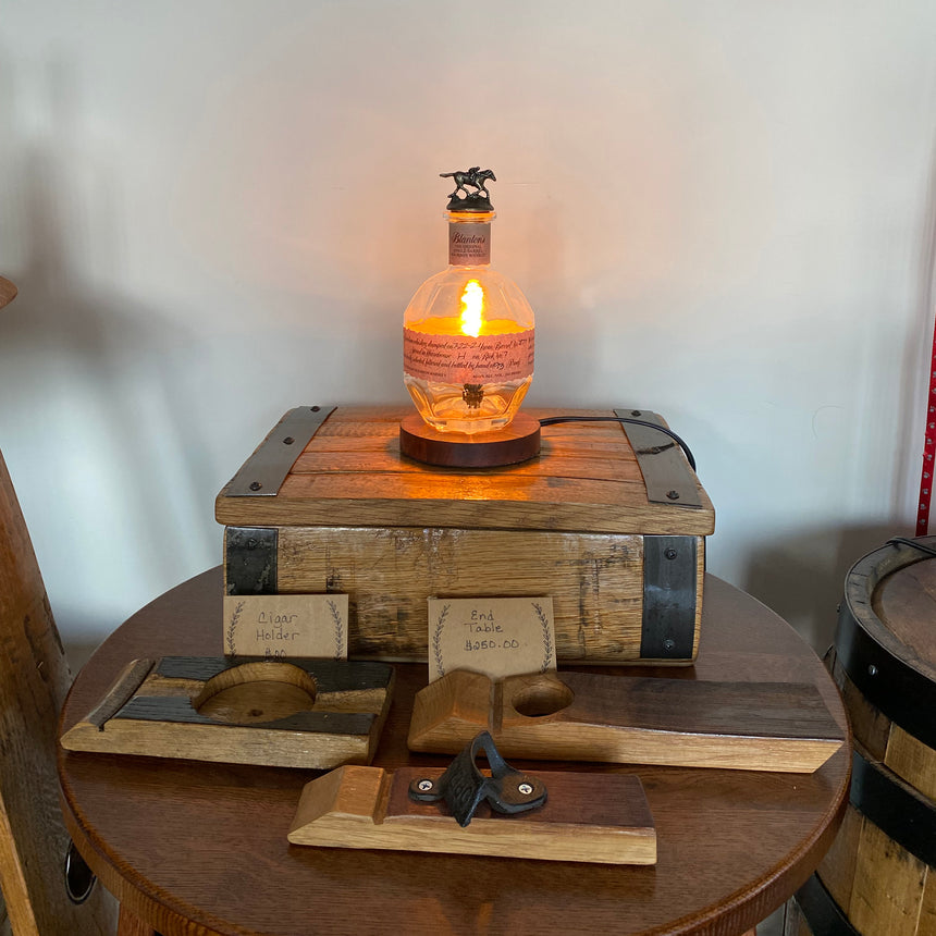 The Blanton's Lamp is an elegant lighting piece featuring a light bulb inside a whiskey bottle set on a wooden chest, with antique hand planes displayed in front.