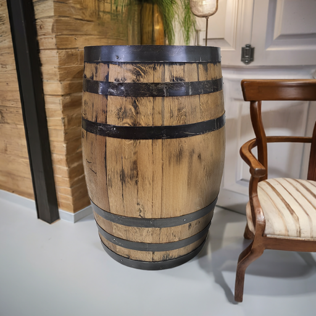 The Standard Refurbished Whiskey Barrel with black metal bands stands beside a wooden chair, adding rustic elegance to the cozy room.