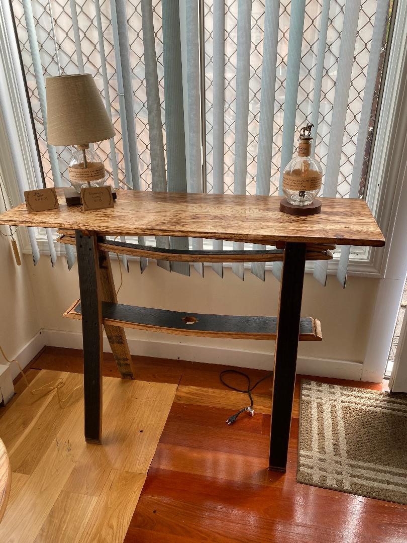Stave Sofa table with a Burnt Hickory Top, a lamp, and decor items, along with a shelf underneath. Placed in front of vertical blinds by the window. Accessories not included. Priced at $325.00.
