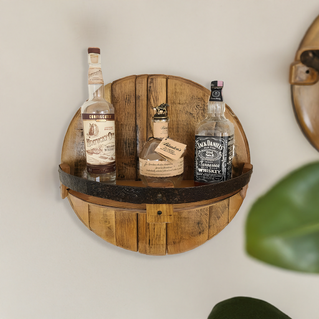 The Whiskey Bottle Shelf, hand made from solid wood barrel construction, stores up to 3 bottles and is shown against a plain wall with a green plant partially visible.
