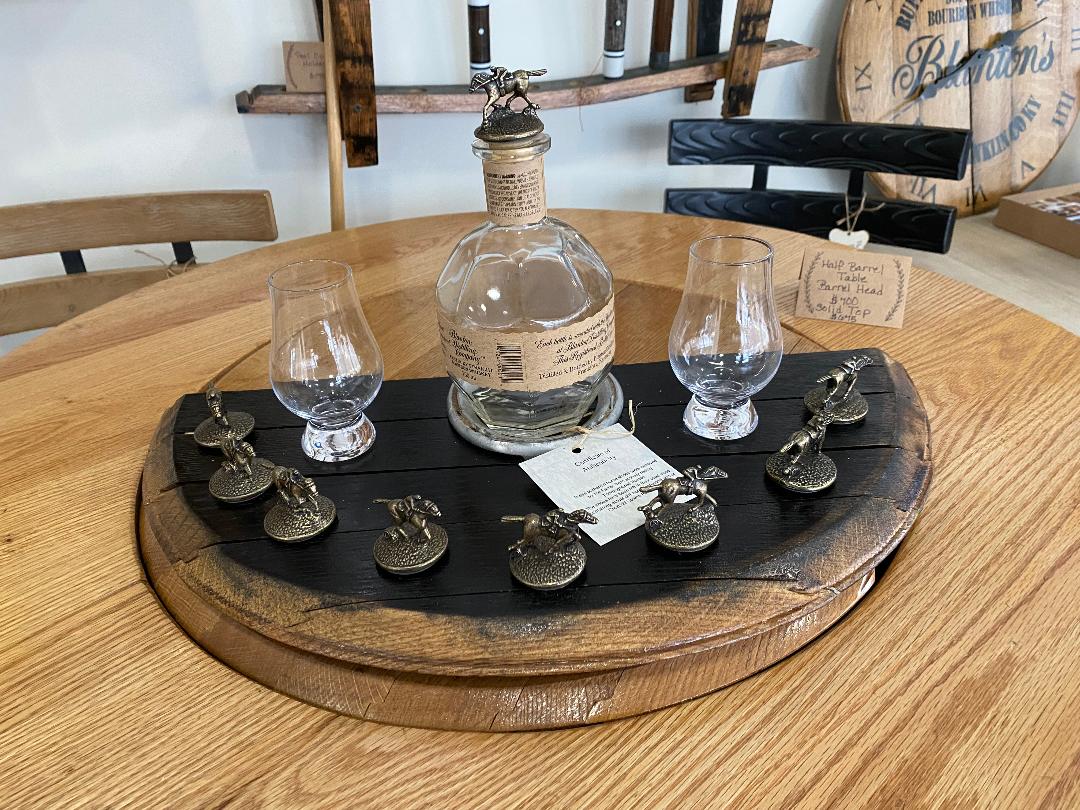 A Whiskey Barrel Head Blanton Bottle and Topper Display priced at $150.00 is set on a table, featuring an empty Blanton bottle, two glasses, and several metal figurines arranged in a semi-circle.