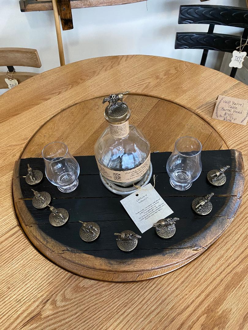 The Whiskey Barrel Head Blanton Bottle and Topper Display, priced at $150.00, features a round wooden tray crafted from a whiskey barrel head that holds a Blanton bottle, two glasses, and several decorative acorn-shaped bottle stoppers arranged around it.