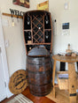 A rustic home bar setup features the Whiskey Barrel Hutch with Wine Rack and Lights ($1750.00), a small wooden table, and wall hooks beneath twinkling lights. The focal point is this charming hutch with its integrated wine rack and welcoming sign on the wall.