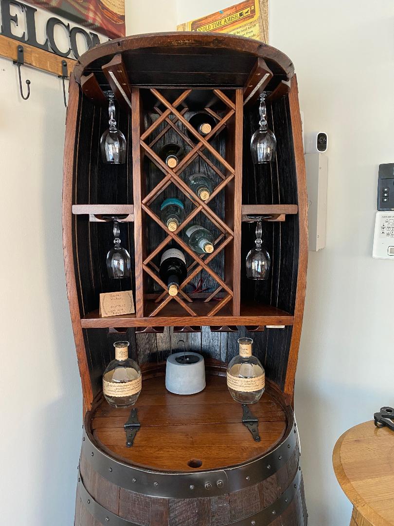 The Whiskey Barrel Hutch with Wine Rack and Lights, priced at $1750.00, boasts a design that cradles wine bottles and glasses alongside two whiskey bottles on a wooden shelf, all softly illuminated by built-in lighting.