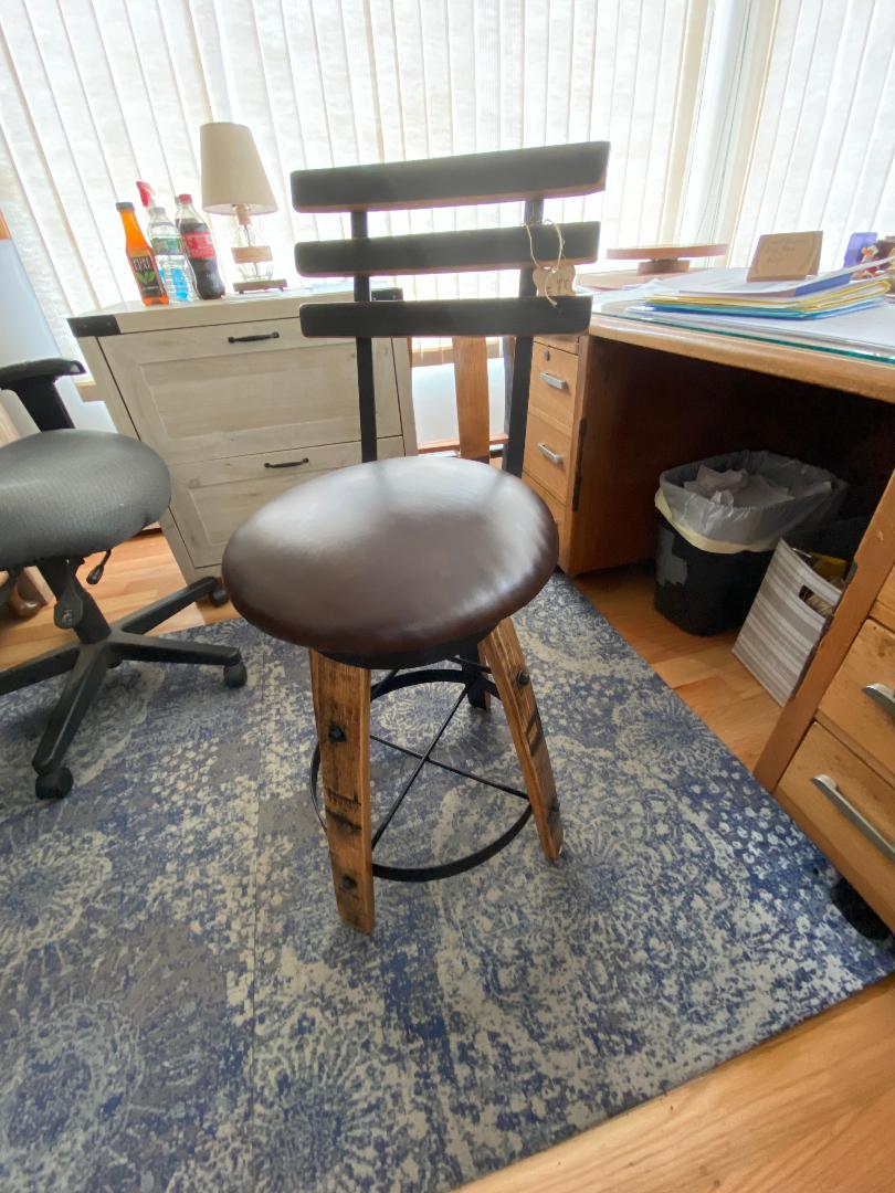 Office space featuring a dark brown cushioned stool, white cabinet, desk, and office chair. A wastebasket sits on a blue rug. Add a touch of uniqueness with our Whiskey Barrel Stave Chair with Back and Cushion for $350.00.