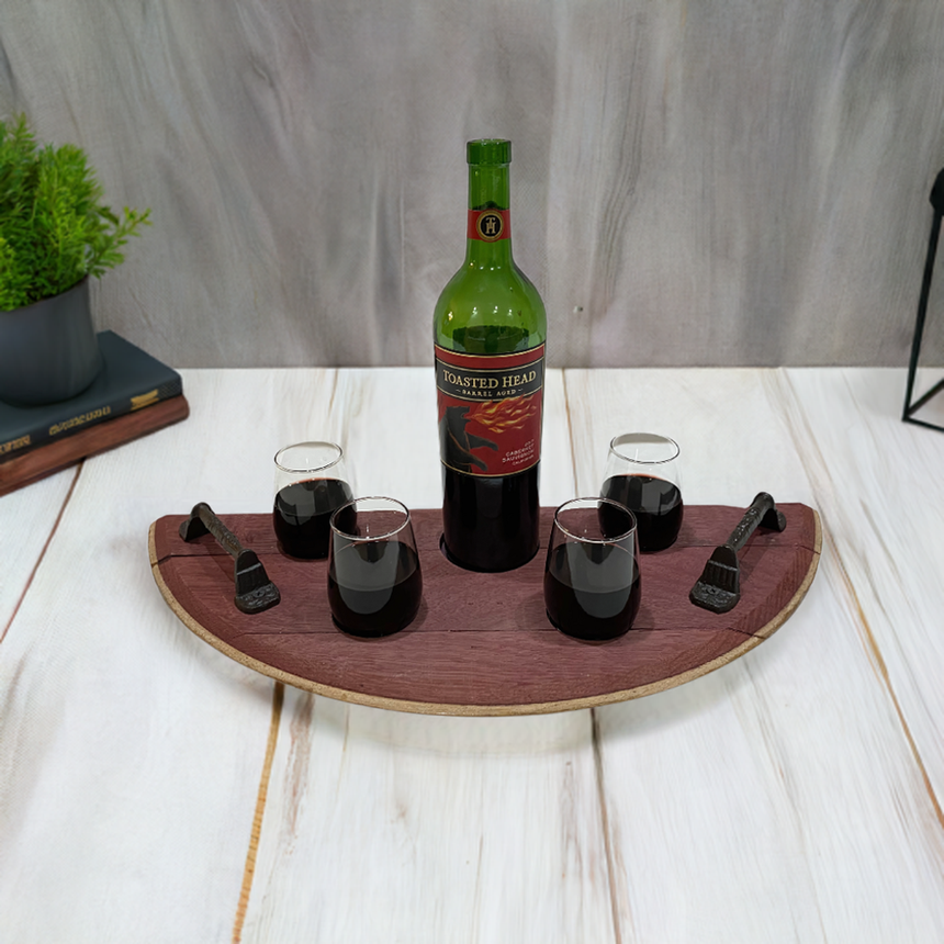 A bottle of red wine and four glasses rest on the Wine Tasting tray - Wine Barrel Head, set on a wooden surface with a green plant and book in the background.
