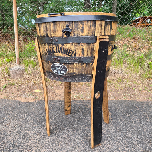 A Whiskey Barrel Cooler with Lid, priced at $395.00 (T008), repurposed into a large planter exudes rustic charm as it stands on a paved surface near a fence.