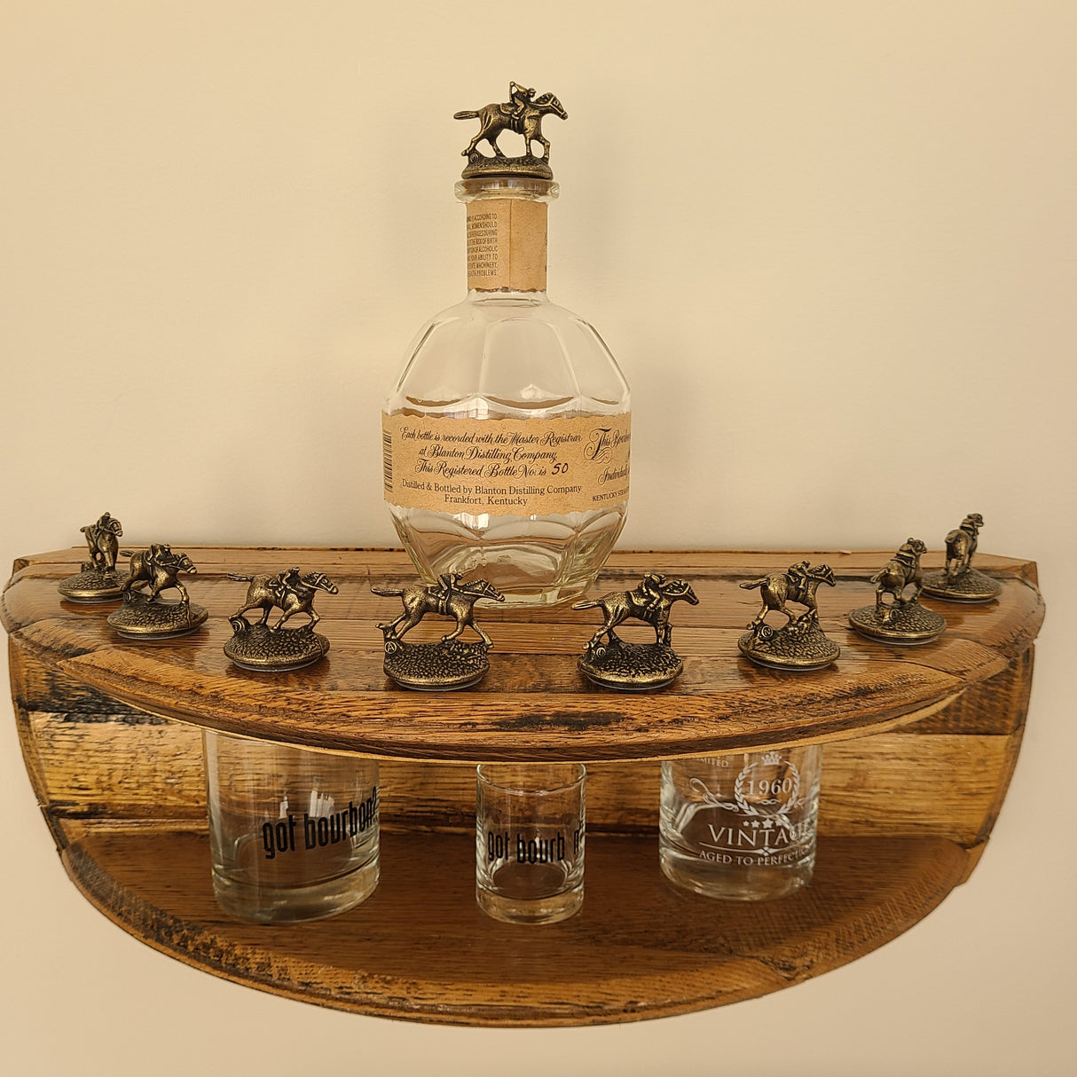 The Blanton's Shelf - A002 ($125.00) features a bottle of bourbon, several collectible lids adorned with horse figurines, and a few glass cups, making it the perfect addition to any whiskey lover’s bourbon collection as it sits elegantly on its wooden construction.