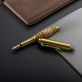 The Bourbon Barrel Fountain Pen, handcrafted from reclaimed Kentucky bourbon barrels with a gold and wood body, rests uncapped on a dark desk beside a notebook.