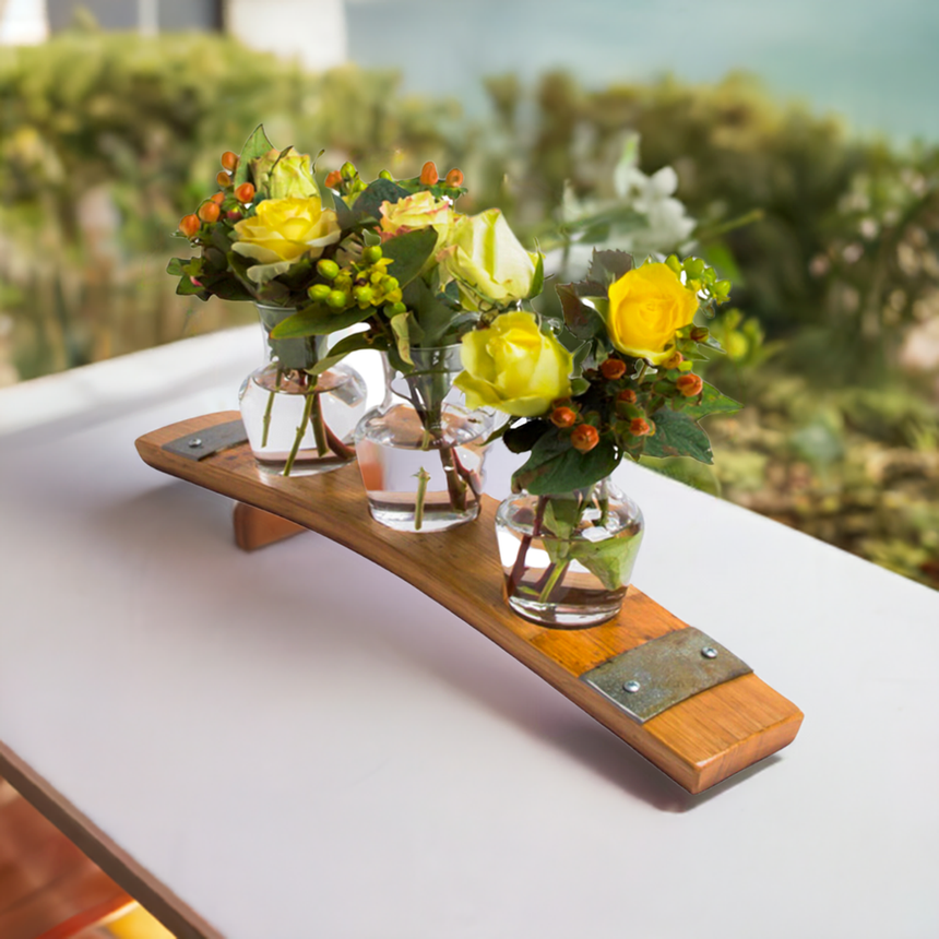 The Bud Vase Stand, crafted from reclaimed whiskey barrels, holds three small vases with yellow roses and greenery, bringing rustic charm to a white table.
