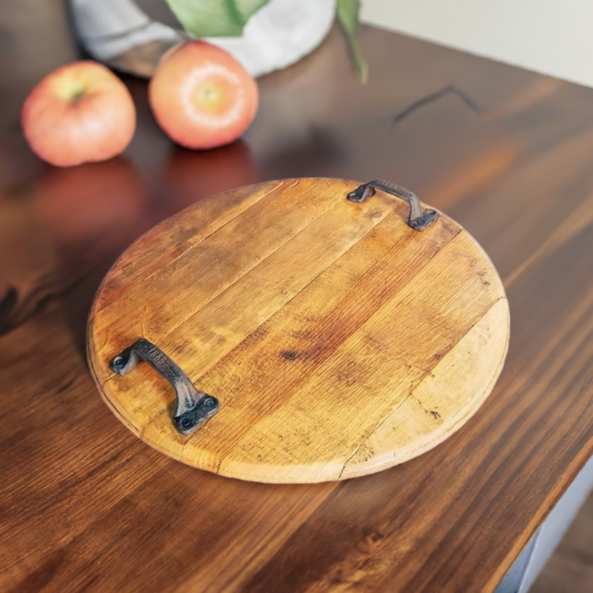 The Charcuterie and Cheese Round Wooden Serving Board, full size with hand made solid wood construction, sits on the table with metal handles, its natural finish echoing whiskey barrels; two apples are in the background.