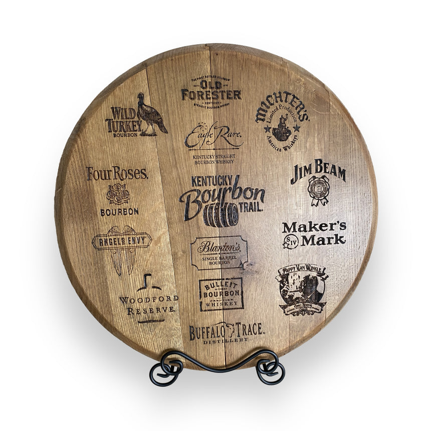The Whiskey Barrel Head Etch Bourbon Trail showcases etched Kentucky bourbon brand logos on a round wooden barrel head, set on a black stand—a unique decor piece inspired by the Bourbon Trail.