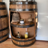 A wooden barrel converted into a shelf featuring bottles and a bag labeled "Bourbon Smoking Chips," placed beside the Deluxe Half Barrel Side Table with Shelves (T001) priced at $375, adds rustic charm to your decor while providing unique storage options.