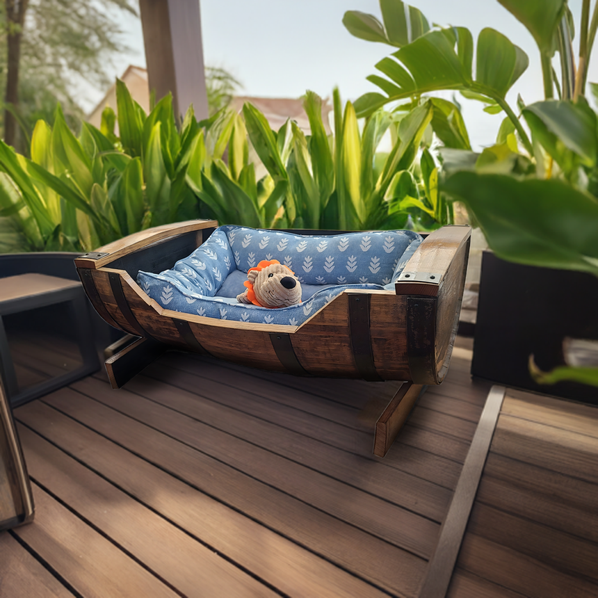 A plush toy lion rests on the Dog Bed Wooden, featuring a handmade solid wood barrel frame and blue cushion, set on a patio surrounded by lush green plants.
