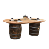 The Double Barrel Table boasts a wooden top supported by two whiskey barrels as legs. It seats four, complete with plates and mugs, and has a central glass-covered display ideal for showcasing barrel furniture pieces.