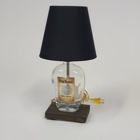 Enhance your home decor with the Four Rose Lamp - A006. Crafted from a Four Roses Small Batch bourbon bottle, this artistic lighting piece features a sleek black shade and elegantly rests on a wooden base. Available for $140.00.