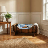 A hand made Dog Bed Wooden with a solid barrel frame and blue cushion sits in a sunny corner by a table with a potted plant, perfect for small to medium dogs.