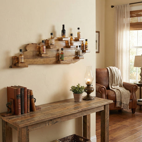 The Kentucky Wood Map Shelf holds liquor bottles on the wall above a rustic table with books, a lamp, plant, and a cozy armchair in the living room.