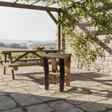 A Bar Stool Wooden, hand made from solid wood barrel staves and available in 24" to 30" heights, brings rustic warmth to a stone patio next to a picnic table, with vines climbing a pergola in the background.