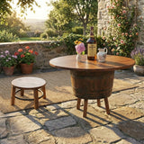 A rustic patio featuring the Barrel Table with Legs, fresh flowers, a whiskey bottle, and a small wooden stool on stone flooring creates timeless charm.
