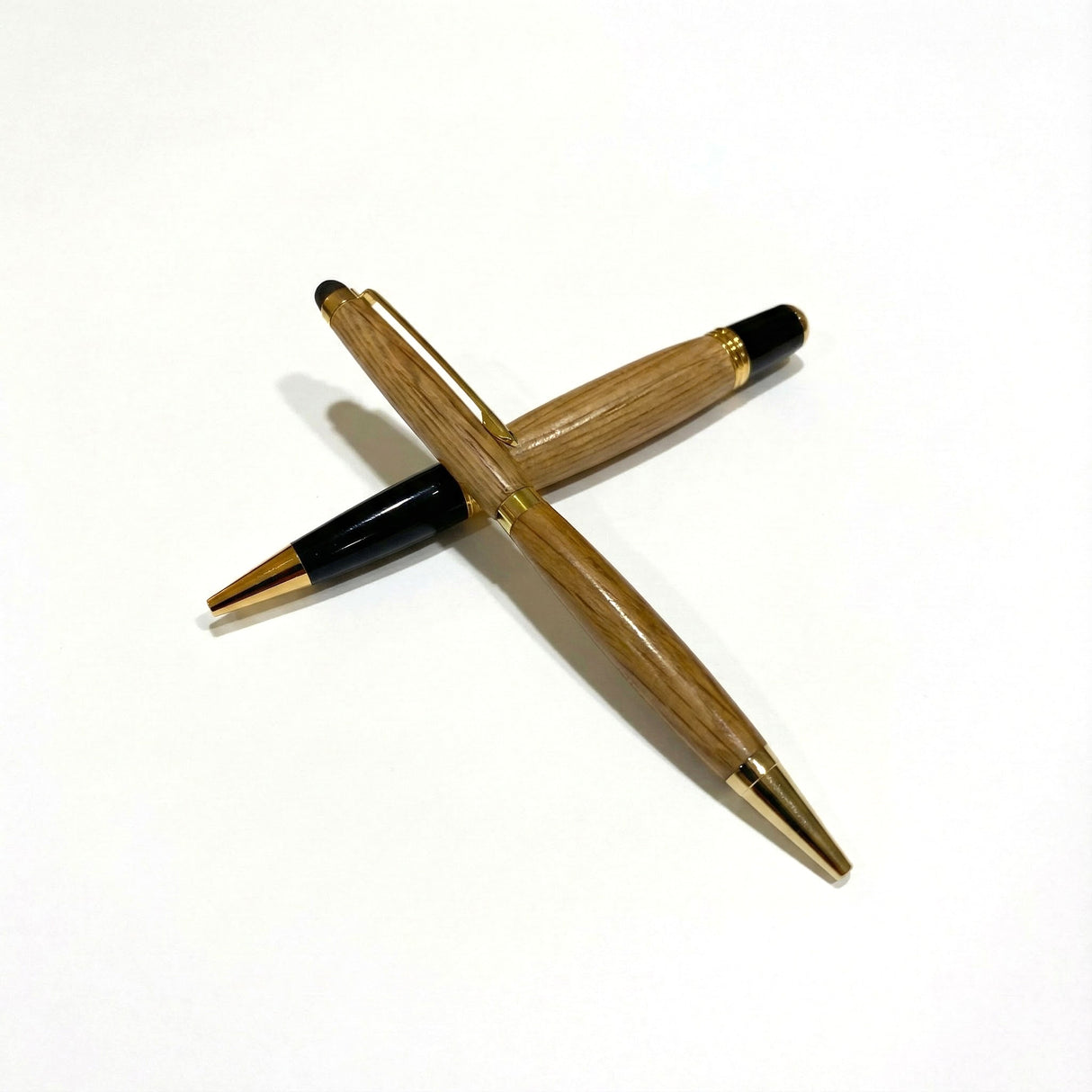 Two Barrel Pens, handcrafted from oak whiskey barrel wood with gold accents, are arranged in a crisscross pattern on a white background.