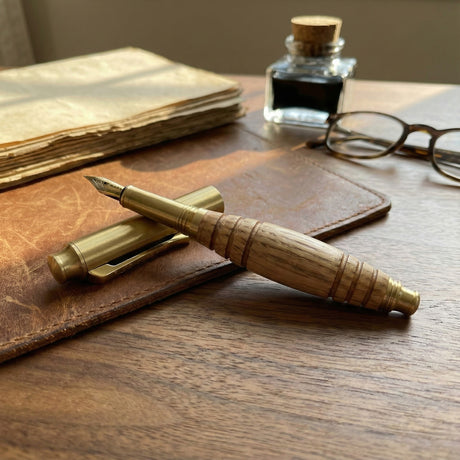 The Bourbon Barrel Fountain Pen, handcrafted from reclaimed Kentucky bourbon barrels, sits on a leather folder next to vintage papers, glasses, and an ink bottle atop a wooden desk.