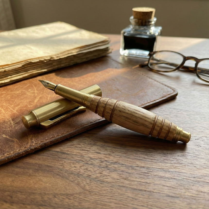 The Bourbon Barrel Fountain Pen, handcrafted from reclaimed Kentucky bourbon barrels, sits on a leather folder next to vintage papers, glasses, and an ink bottle atop a wooden desk.