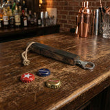 A Bottle Opener with a wooden handle sits next to three bottle caps on a bar counter, with drinks softly blurred in the background.