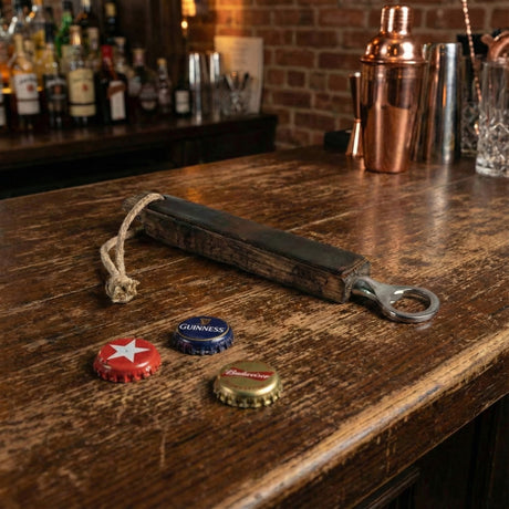 A Bottle Opener with a wooden handle sits next to three bottle caps on a bar counter, with drinks softly blurred in the background.