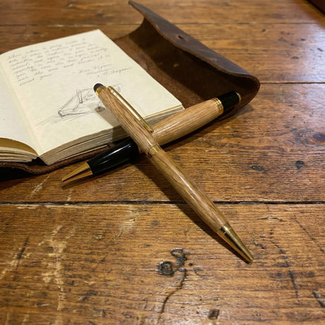 Two handcrafted Barrel Pens crossed on a rustic wooden table beside an open sketchbook and a brown leather case.