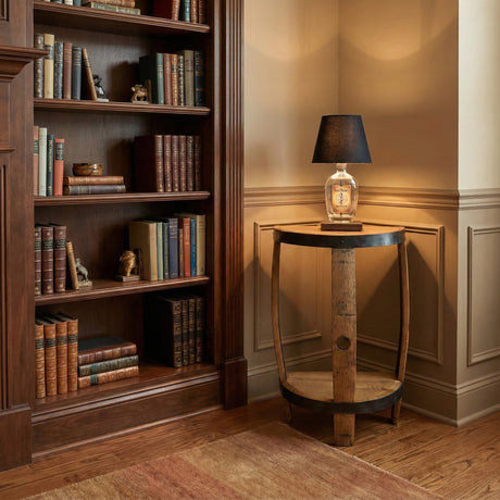 A Lamp Side Table crafted from reclaimed whiskey barrels stands in a warmly lit room, beside a wooden bookshelf and books.