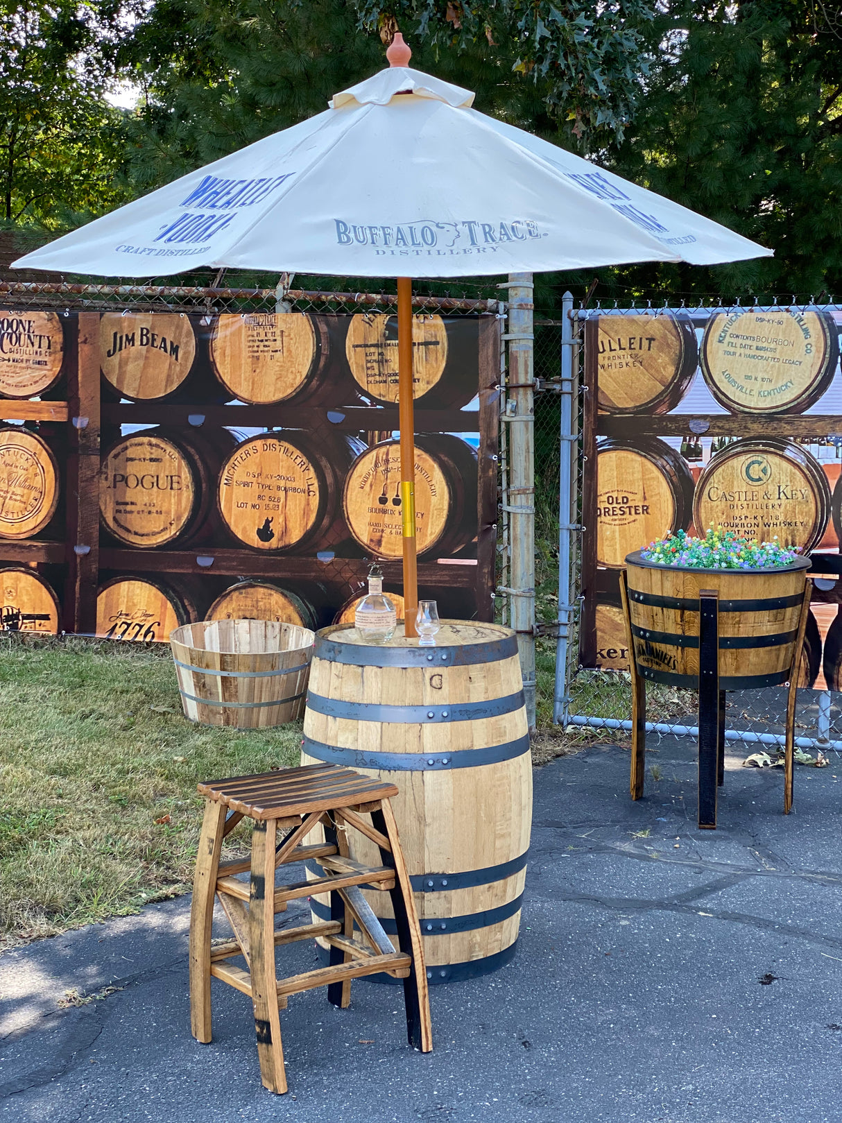 The Patio Pub Table is complemented by an umbrella, chairs, whiskey barrel decor, and bottles on a small table to create the perfect outdoor entertainment setting.