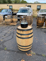 The Pub Table Glass Fire Top sits on whiskey barrels resting on the pavement, adding rustic charm. In the background, parked trucks enhance the industrial vibe.