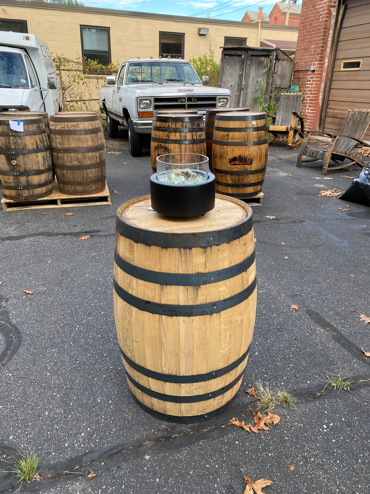 A Pub Table Glass Fire Top resembling a wooden whiskey barrel with a small black dish sits among barrels and parked trucks in a lot, looking like an outdoor fire pit table.