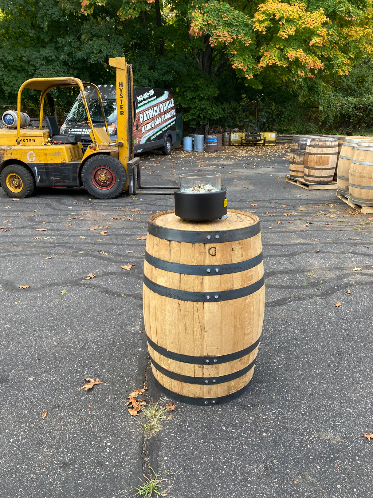 A Pub Table Glass Fire Top sits on a wooden whiskey barrel, mimicking an outdoor fire pit table, surrounded by more barrels and a parked forklift.