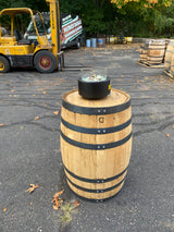 A Pub Table Glass Fire Top sits atop a rustic wooden barrel reminiscent of whiskey barrels, with a forklift and more barrels on the asphalt in the background.