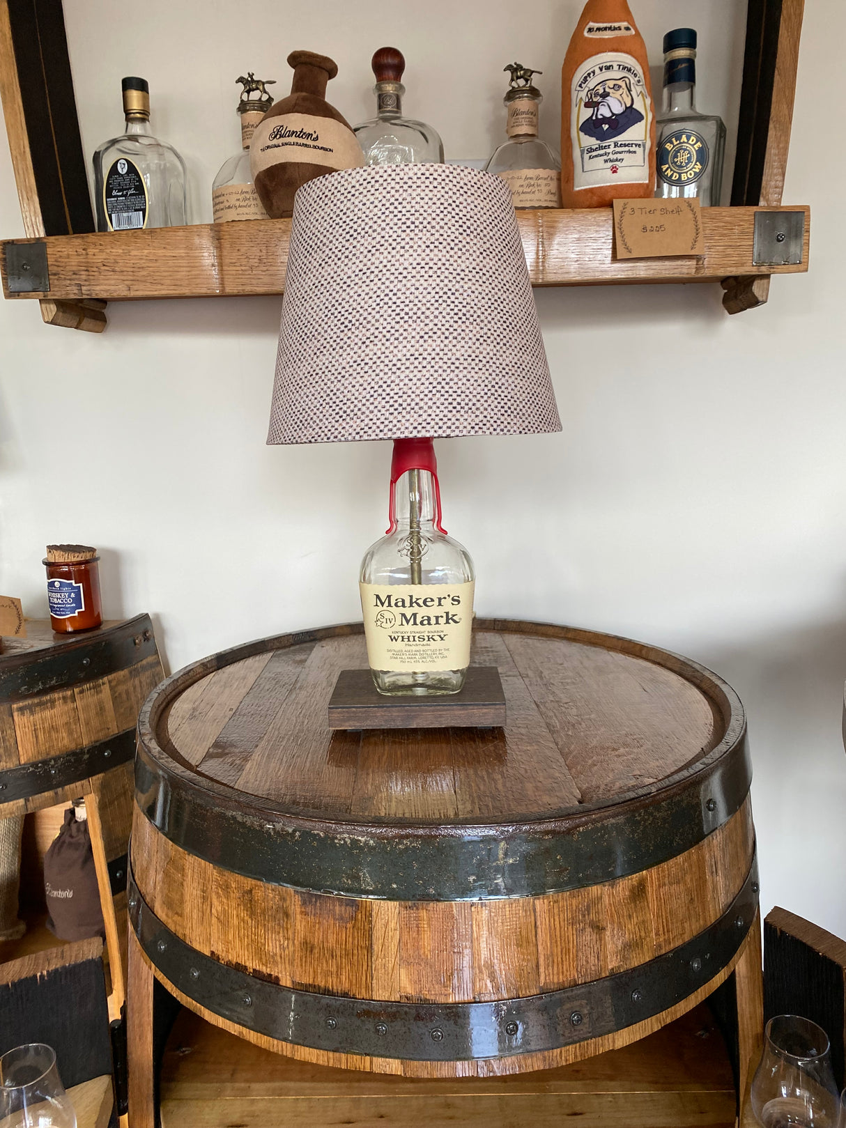 The Maker's Mark Lamp, crafted from a bourbon bottle, sits on a wooden barrel surrounded by whiskey bottles on shelves, making it an ideal home decor piece for enthusiasts.