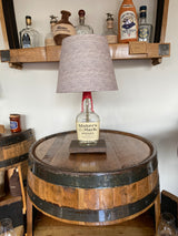 The Maker's Mark Lamp, crafted from a bourbon bottle, sits on a wooden barrel surrounded by whiskey bottles on shelves, making it an ideal home decor piece for enthusiasts.