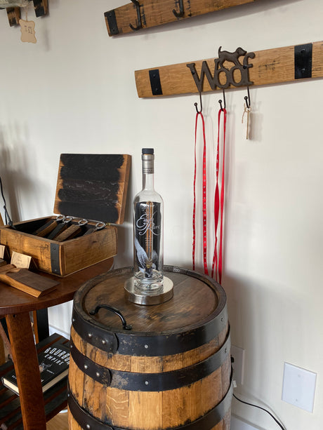 An Eagle Rare Lamp casts a warm glow on a vodka bottle resting on a wooden barrel. Beside it, a vintage wall rack labeled "Woof" with hanging leashes completes the scene, adding rustic charm to the cozy nook.